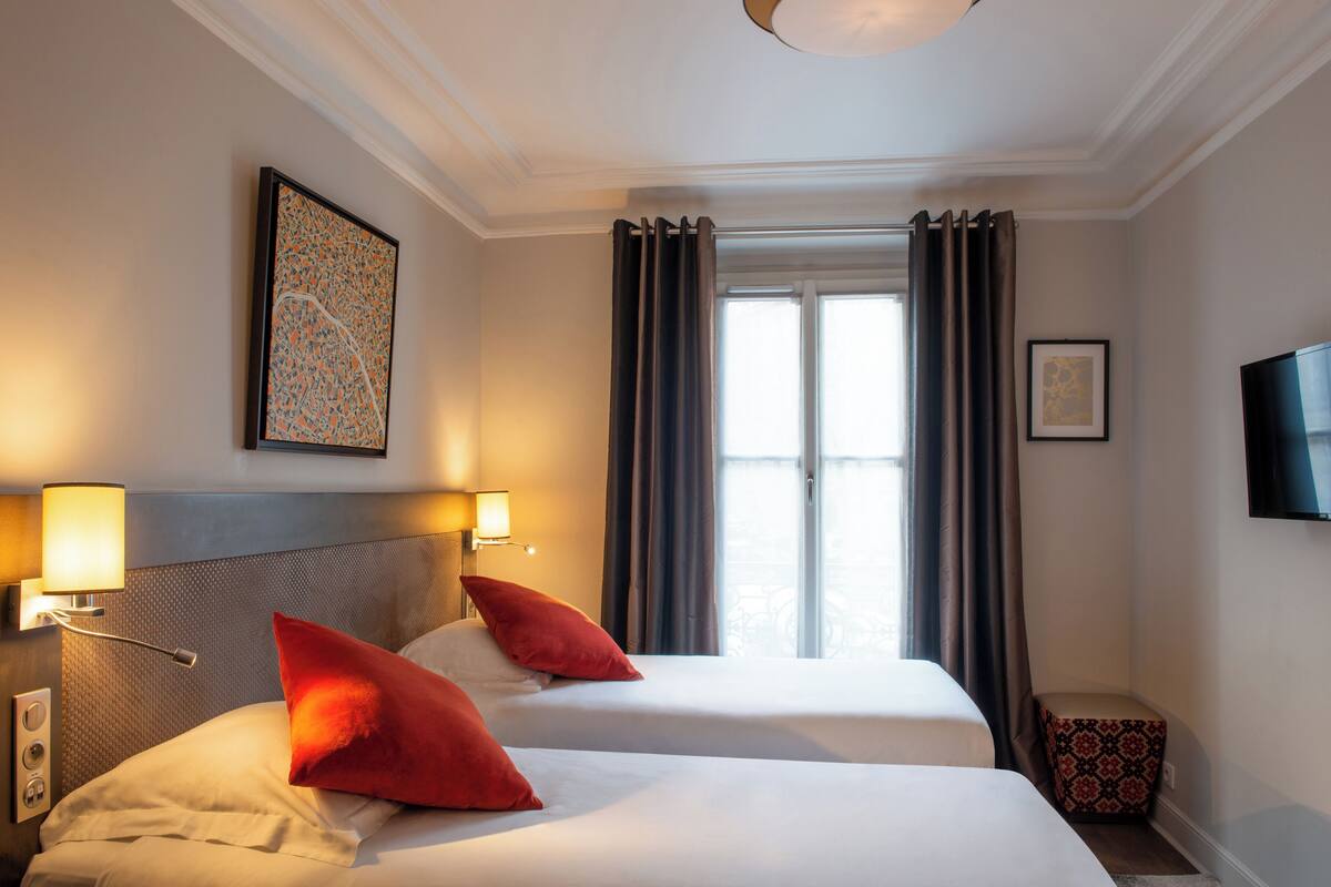 A hotel room with two beds, a large window with curtains, a flat-screen TV, and wall-mounted lamps.
