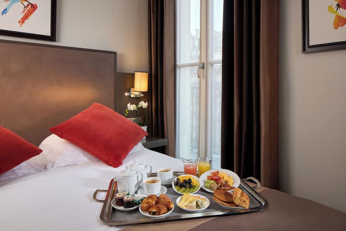 A neatly made bed with red pillows, a tray of breakfast including croissants, fruit, and pastries, and a view of the city through a window.