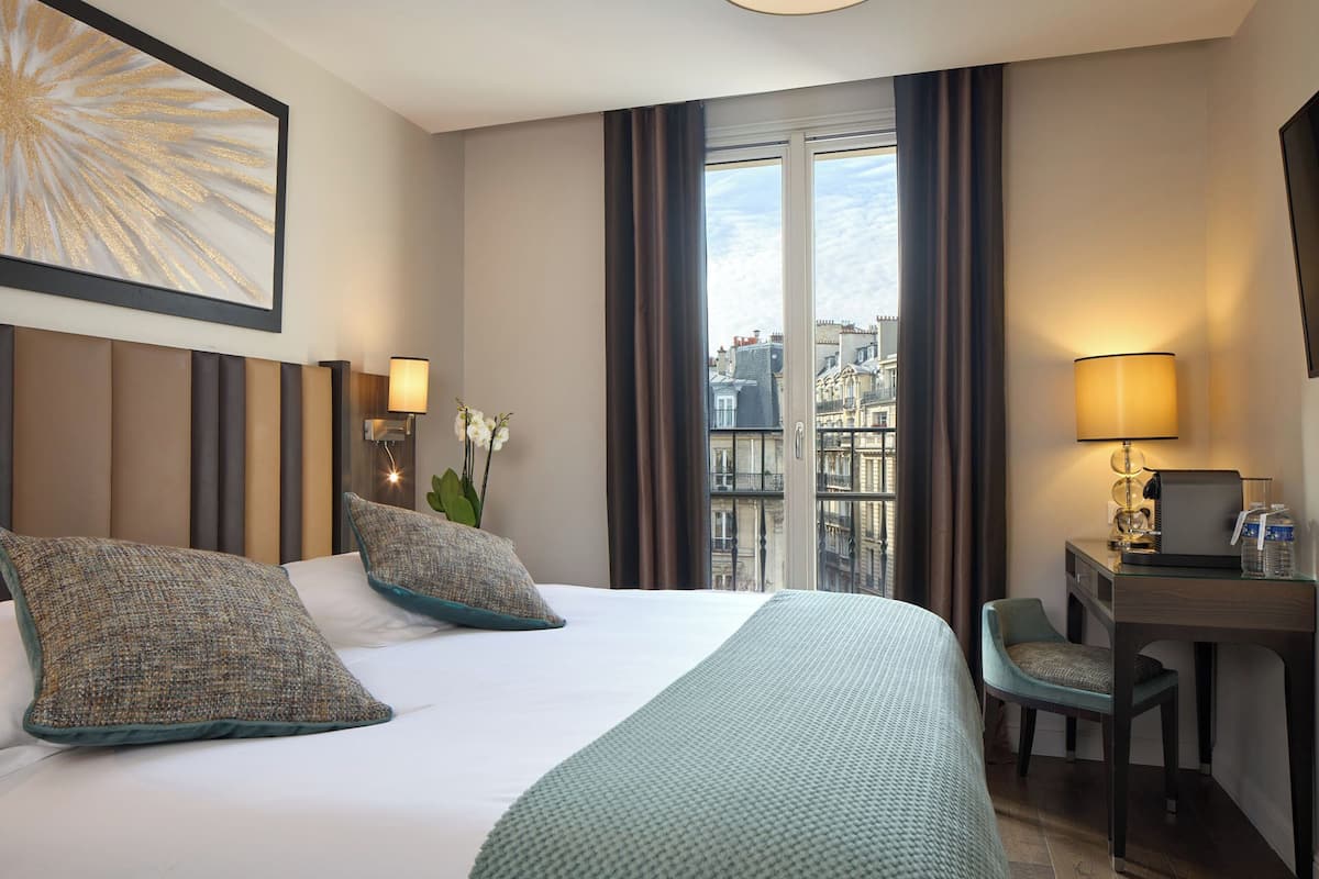 A hotel room with a large bed, two pillows, a desk with a chair, a lamp, and a view of buildings through a window.