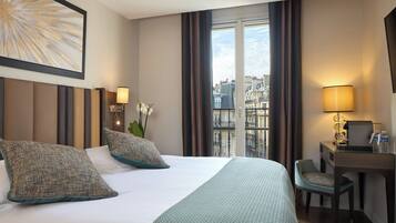 A hotel room with a large bed, two pillows, a desk with a chair, a lamp, and a view of buildings through a window.