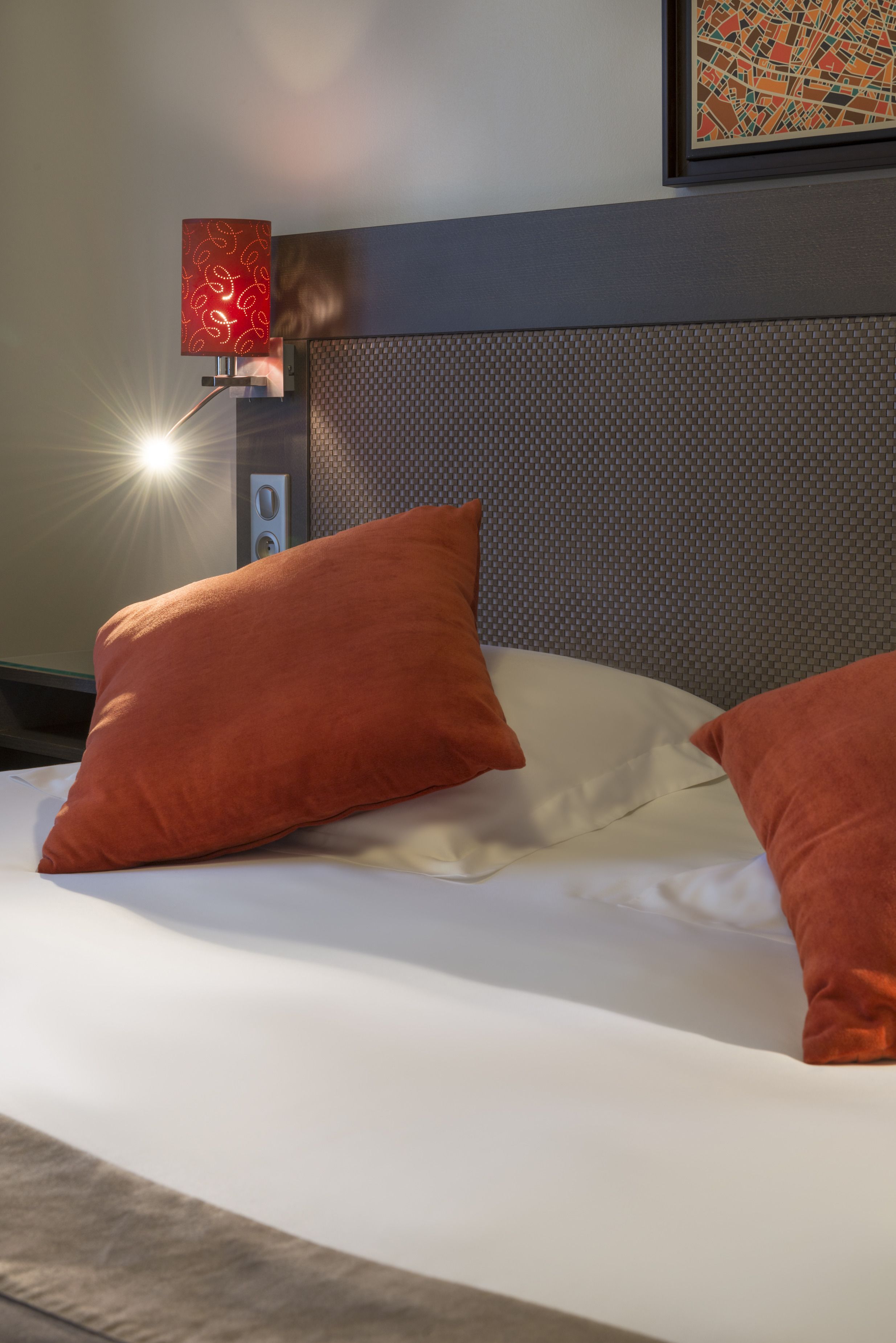 Habitació d'hotel amb un llit, dos coixins taronja, un capçal de llit i una làmpada penjada a la paret.