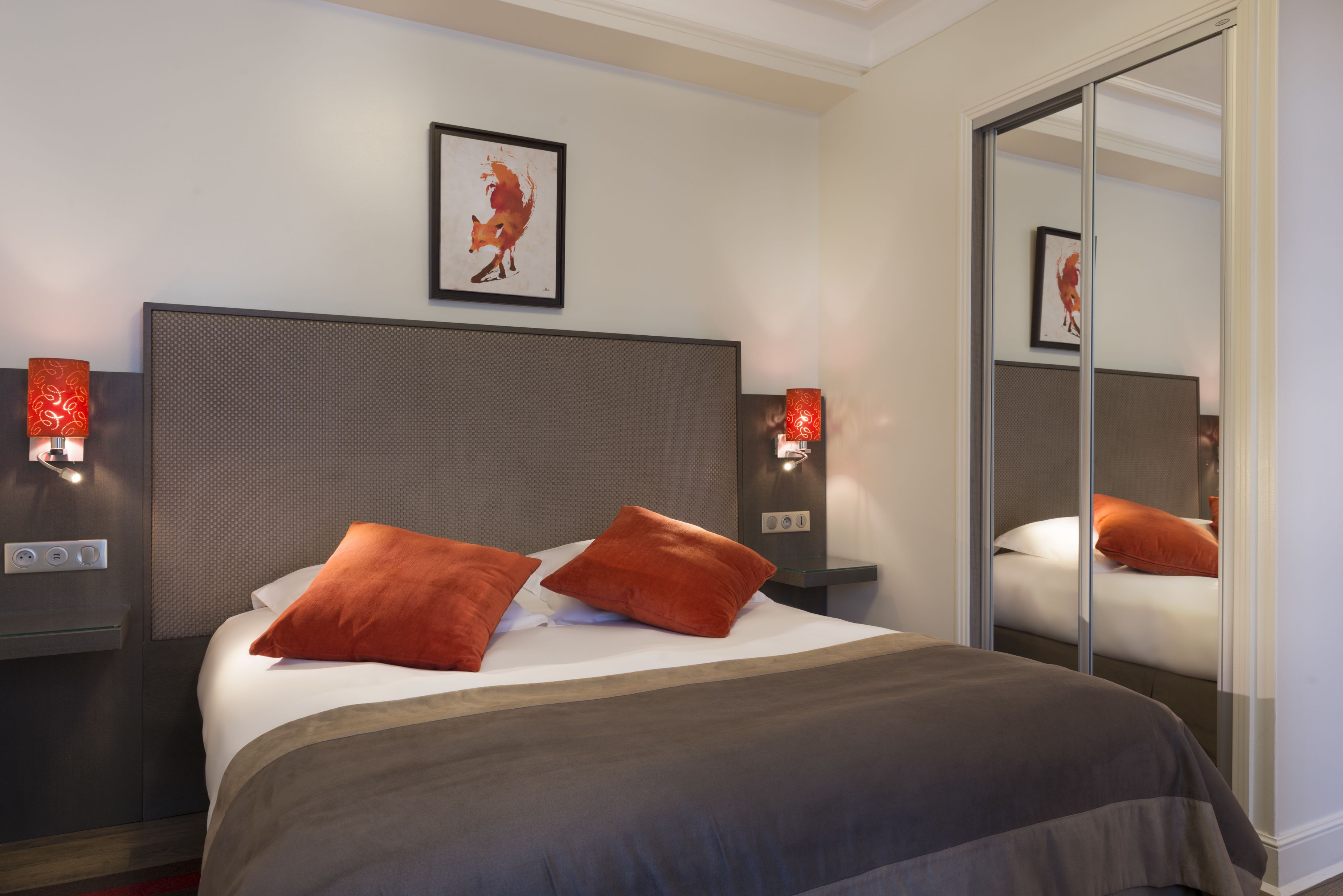 A hotel room with a large bed, two red pillows, a headboard with a textured design, a framed picture on the wall, and a mirror reflecting the room.