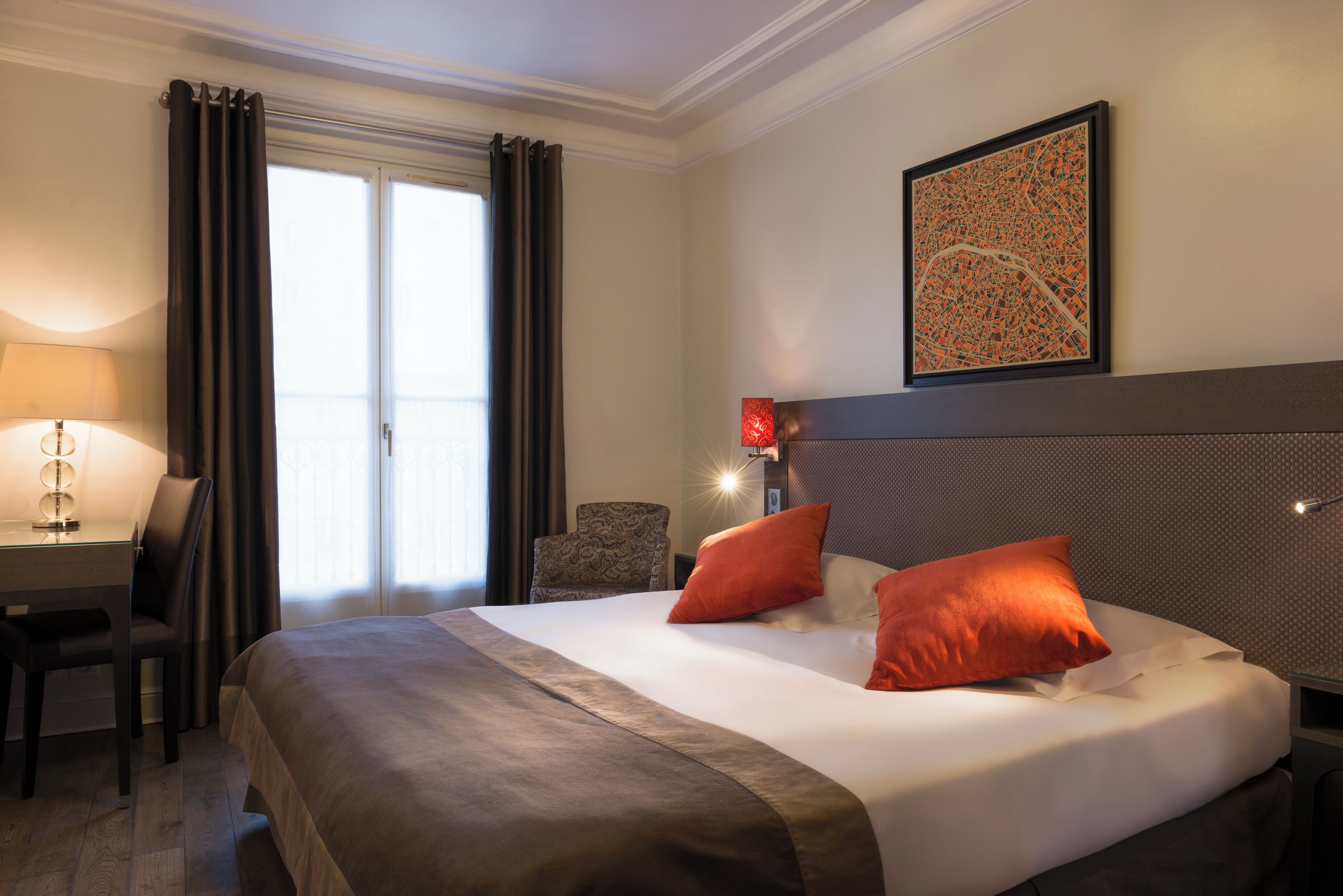 A hotel room with a large bed, two pillows, a desk with a lamp, a chair, and a window with curtains.