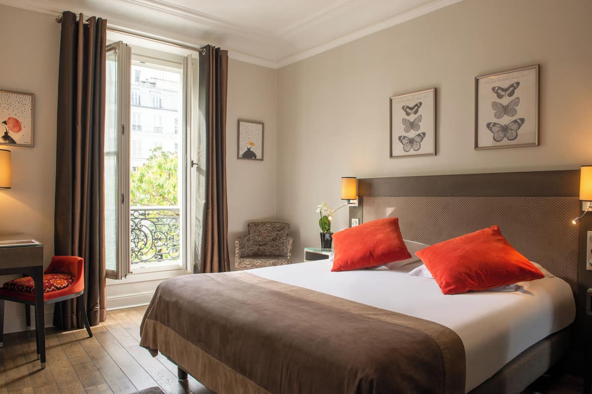 A hotel room with a large bed, two red pillows, a chair, a desk, and a window with curtains.