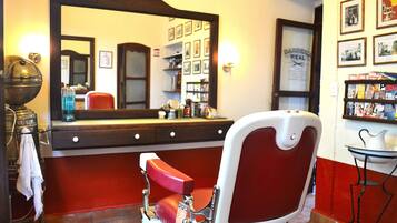 Un sillón de barbero con cojín rojo, un espejo grande y una estantería con revistas y artículos de aseo.