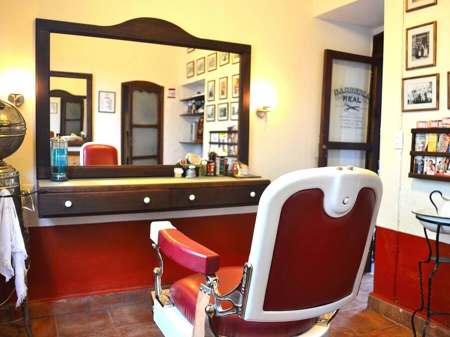 Un sillón de barbero con cojín rojo, un espejo grande y una estantería con revistas y artículos de aseo.
