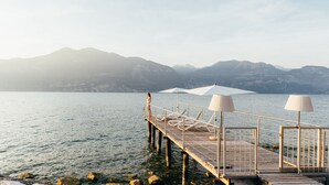 Un muelle de madera se extiende sobre aguas tranquilas, con una montaña al fondo. Hay sillas de descanso y sombrillas en el muelle.