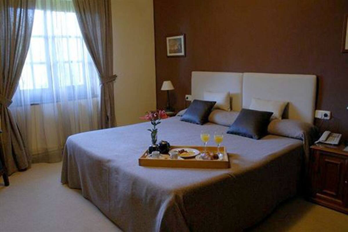 Habitación de hotel con una cama grande, una bandeja con alimentos para el desayuno, una mesita de noche y una ventana con cortinas.