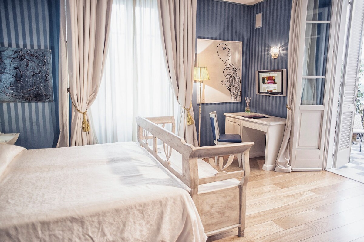 A bedroom with a bed, a desk, and a chair. There is a window with curtains, a lamp, and artwork on the wall.
