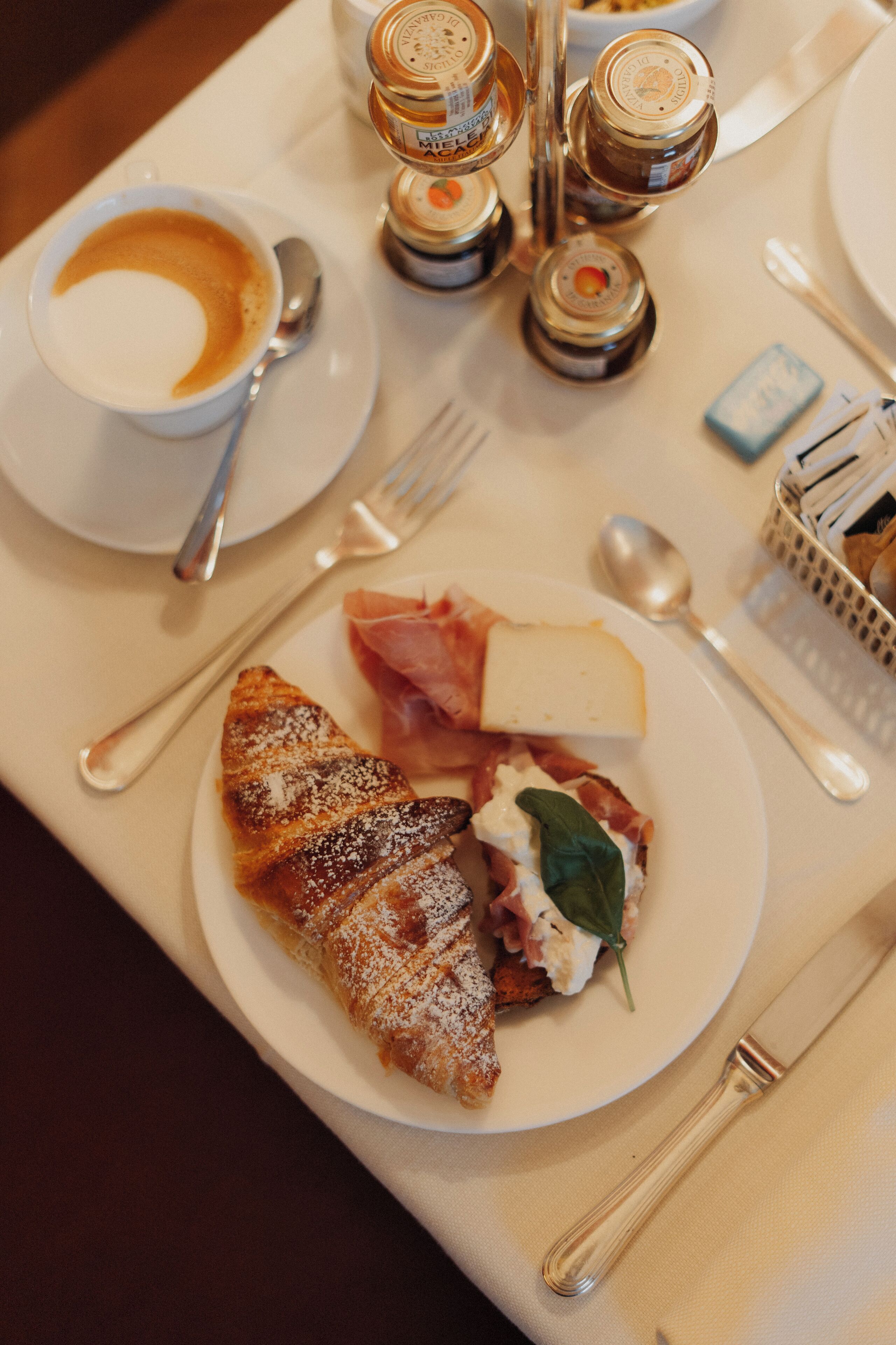 A breakfast setting with a croissant, cheese, and ham on a plate, a cup of coffee, and condiments on the table.