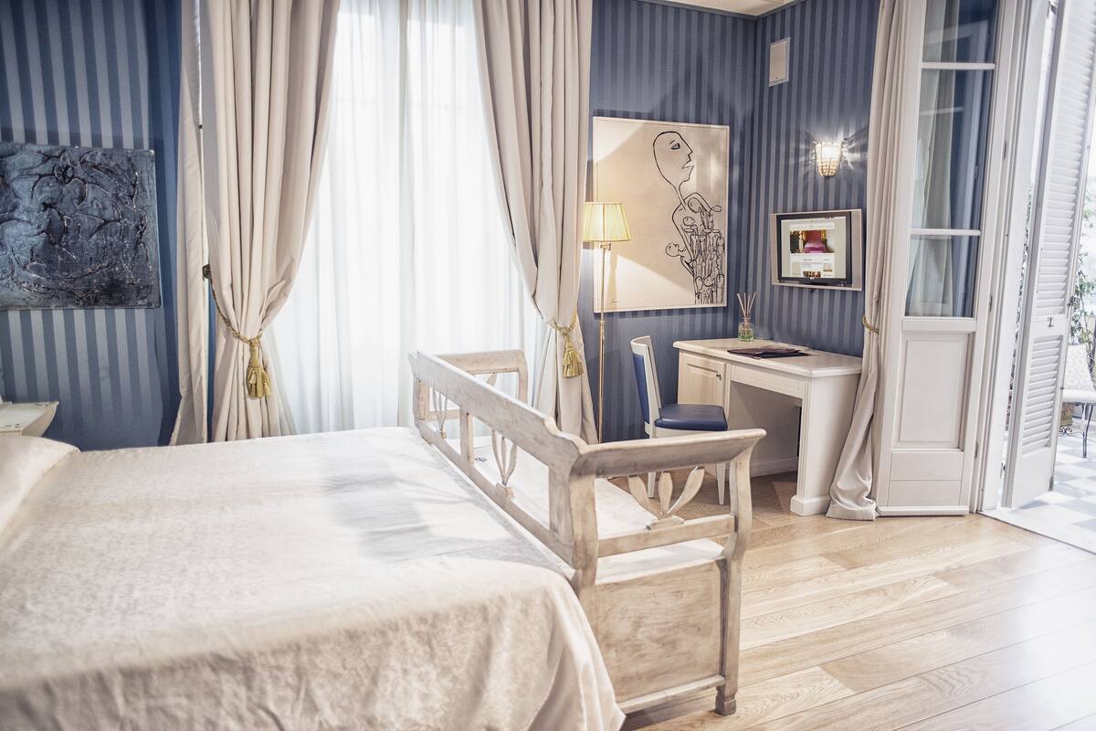 A bedroom with a bed, a desk, and a chair. There is a window with curtains, a lamp, and artwork on the wall.