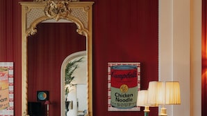 A living room with a patterned sofa, a red ottoman, a wooden coffee table, a lamp, a bookshelf, and a large mirror. - Hotel Palazzo Guiscardo (Pietrasanta)