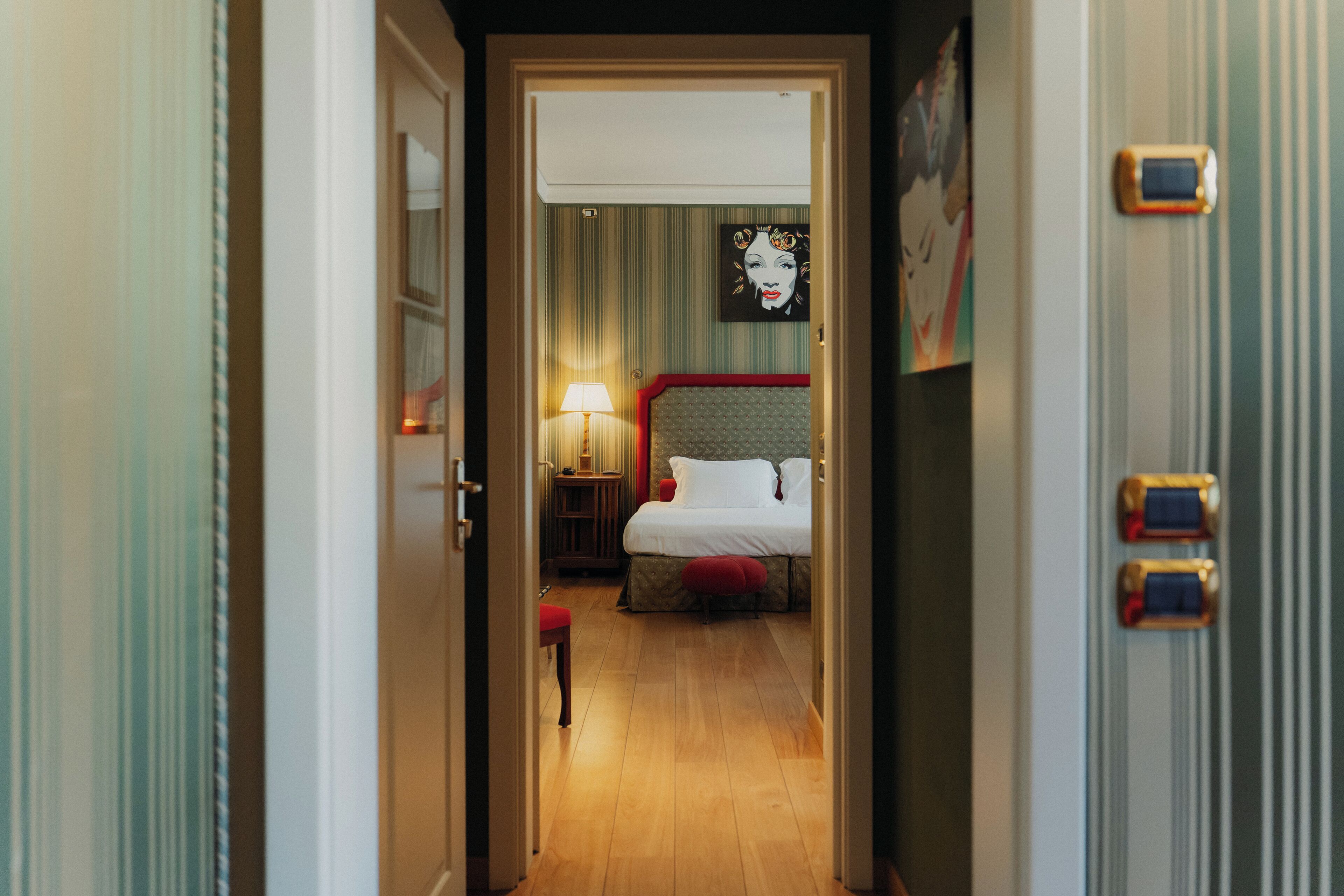Una habitación de hotel con una cama, una mesita de noche, una lámpara y un taburete rojo.