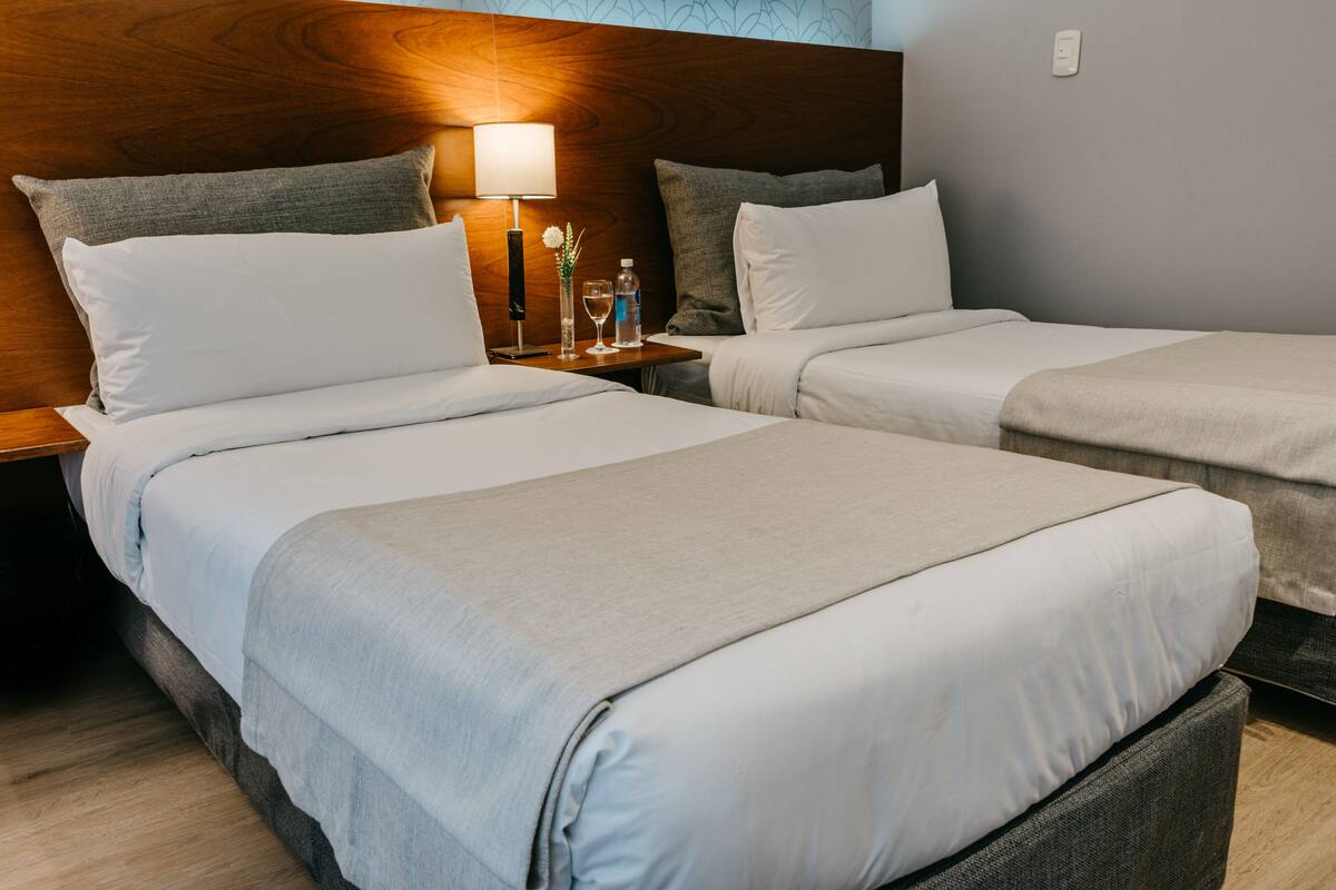 A hotel room with two single beds, a wooden headboard, a nightstand with a lamp, a vase with flowers, and a bottle of water.