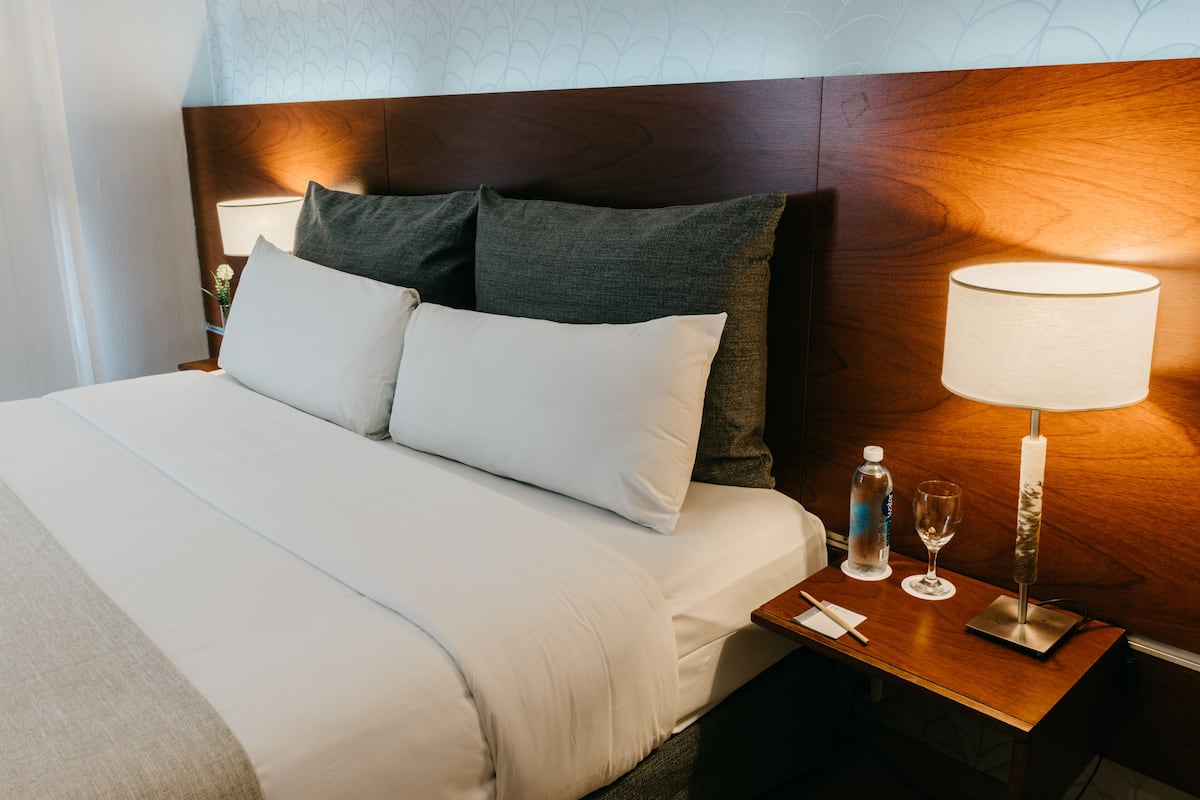A neatly made bed with white linens, a wooden headboard, and a bedside table with a lamp, water bottle, and glass.
