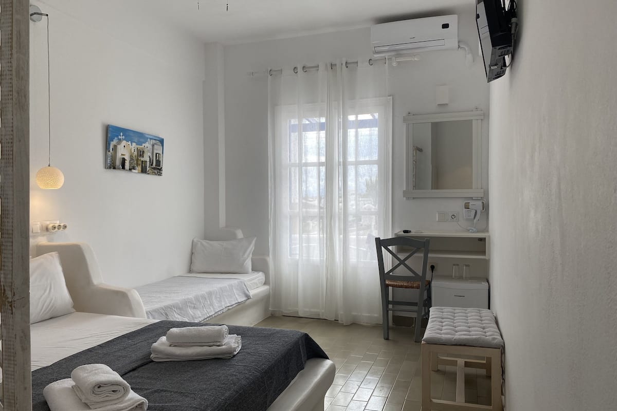 A hotel room with two single beds, a desk with a chair, a small table, a mirror, and an air conditioning unit.