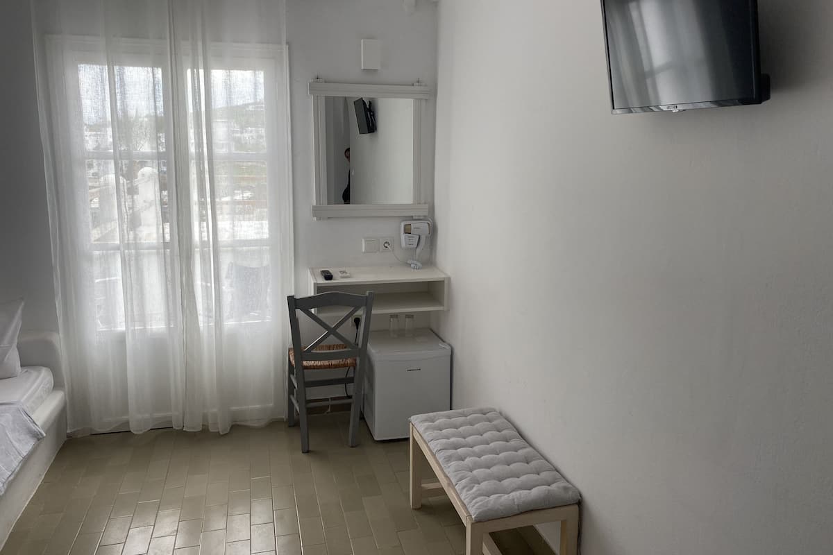 A small, clean room with a bed, a desk, a chair, a bench, a TV, and a window with curtains.
