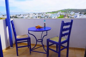 Een balkon met twee blauwe stoelen en een klein tafeltje, met uitzicht op een kustplaats met witte gebouwen.