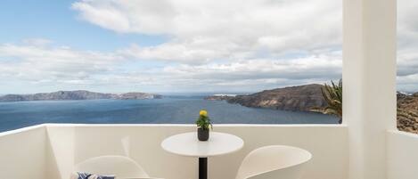 Een balkon met een witte tafel en twee witte stoelen, een bloempot en uitzicht op zee en heuvels.