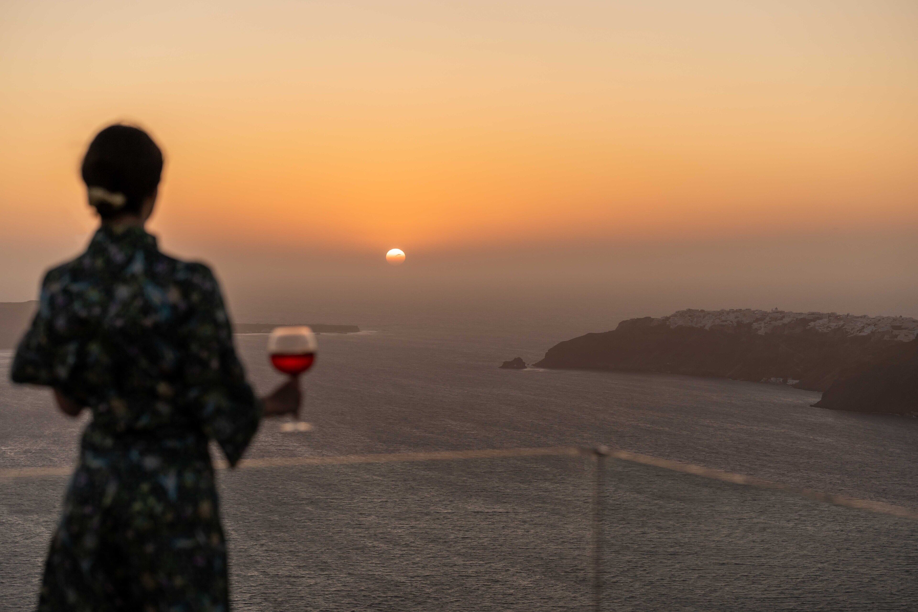 A person in a patterned dress standing by the sea, holding a glass of red wine, with a sunset in the background.