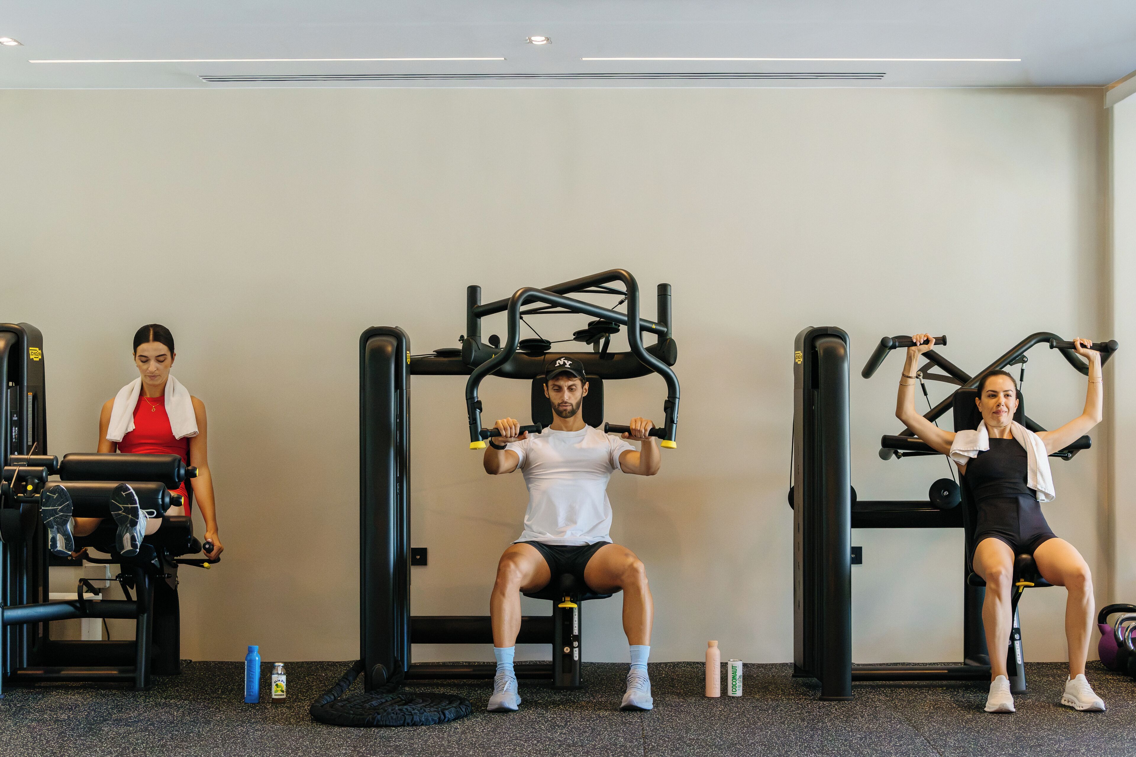 Tres personas haciendo ejercicio en máquinas de gimnasio distintas en un espacio con piso moteado y pared lisa.