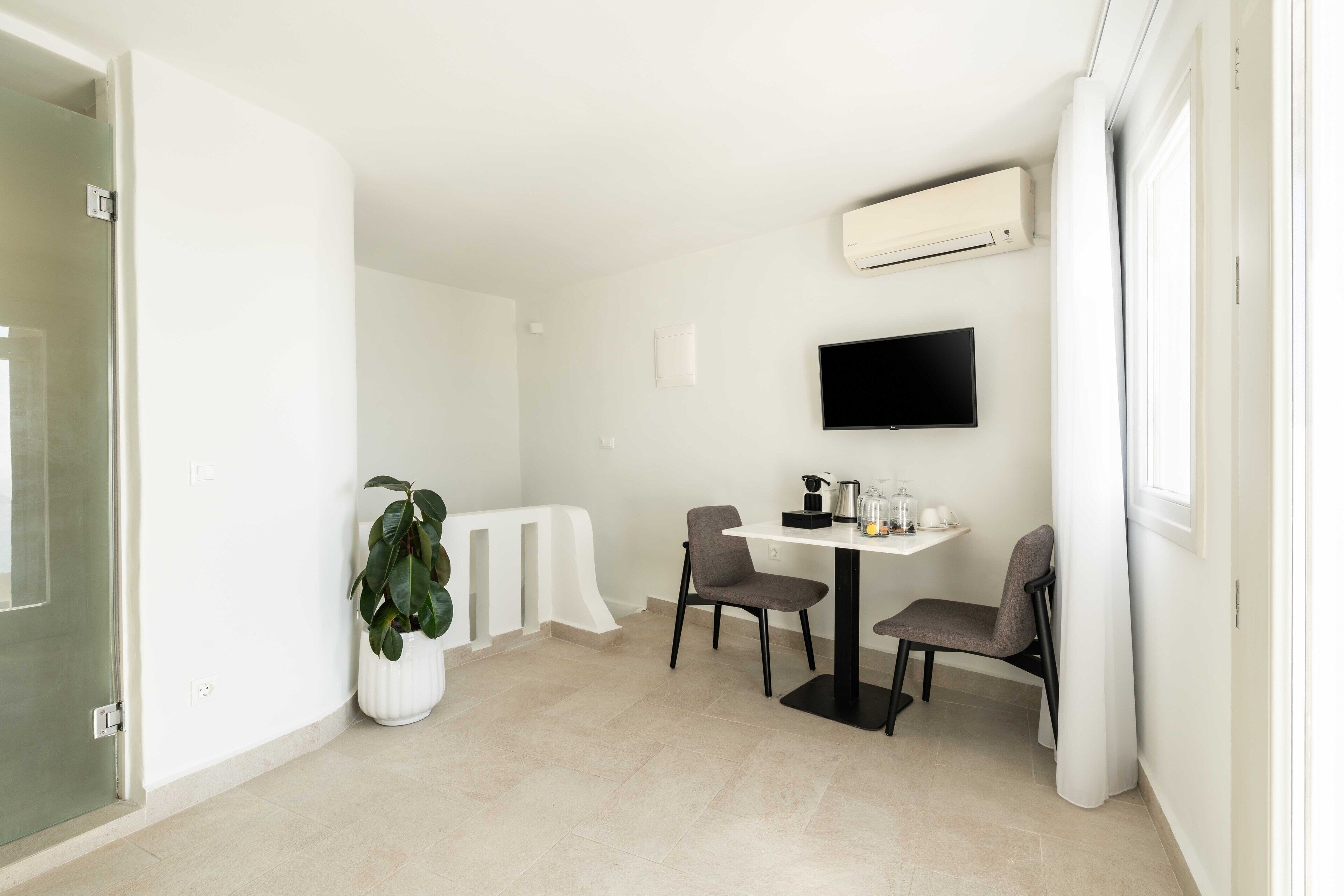 Una habitación moderna y luminosa con una mesa de comedor, dos sillas, una planta en maceta, un televisor de pantalla plana montado en la pared y un equipo de aire acondicionado.