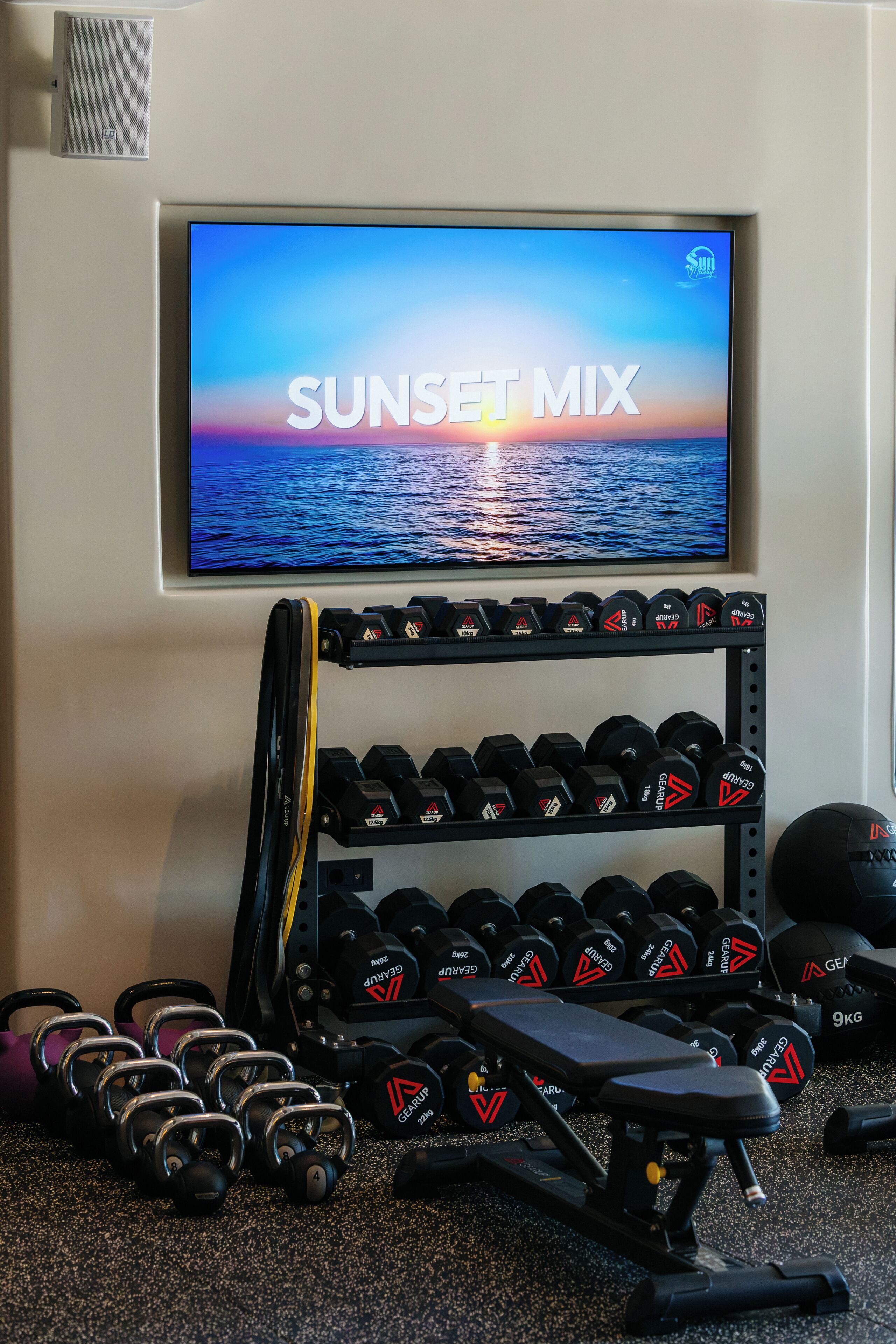 Un gimnasio en casa con una televisión mostrando 'SUNSET MIX', un estante de mancuernas y una banca.