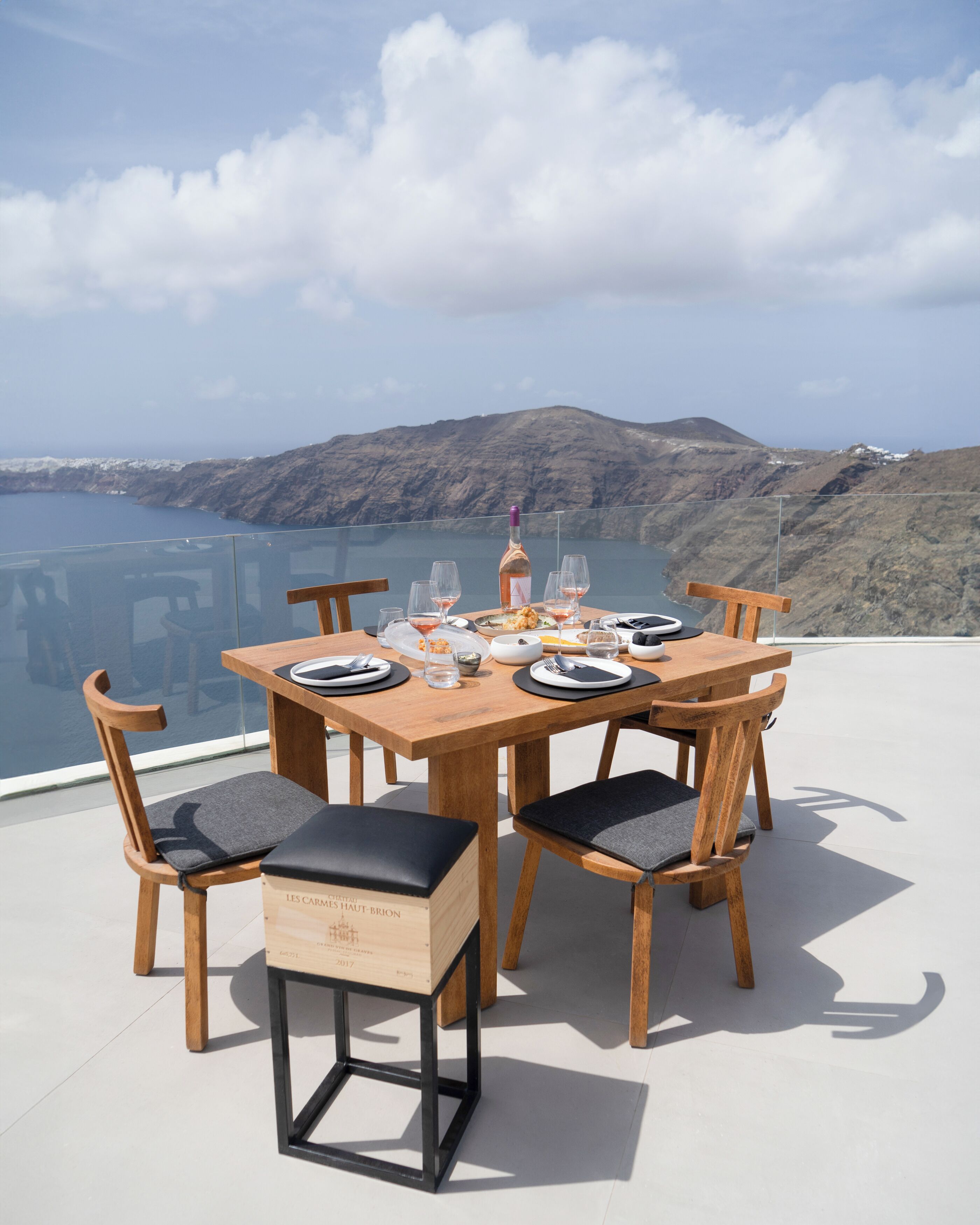 A wooden dining table set with plates, glasses, and a bottle, overlooking a scenic coastal landscape.