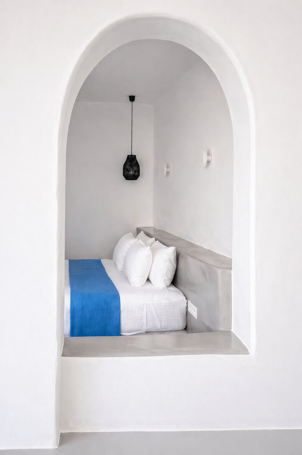 A small, white, arched alcove with a bed, a blue blanket, and a hanging light fixture.