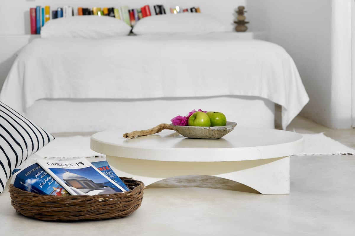 A neatly arranged bedroom with a bed, a round coffee table, a wicker basket with magazines, and a bowl with apples and flowers.