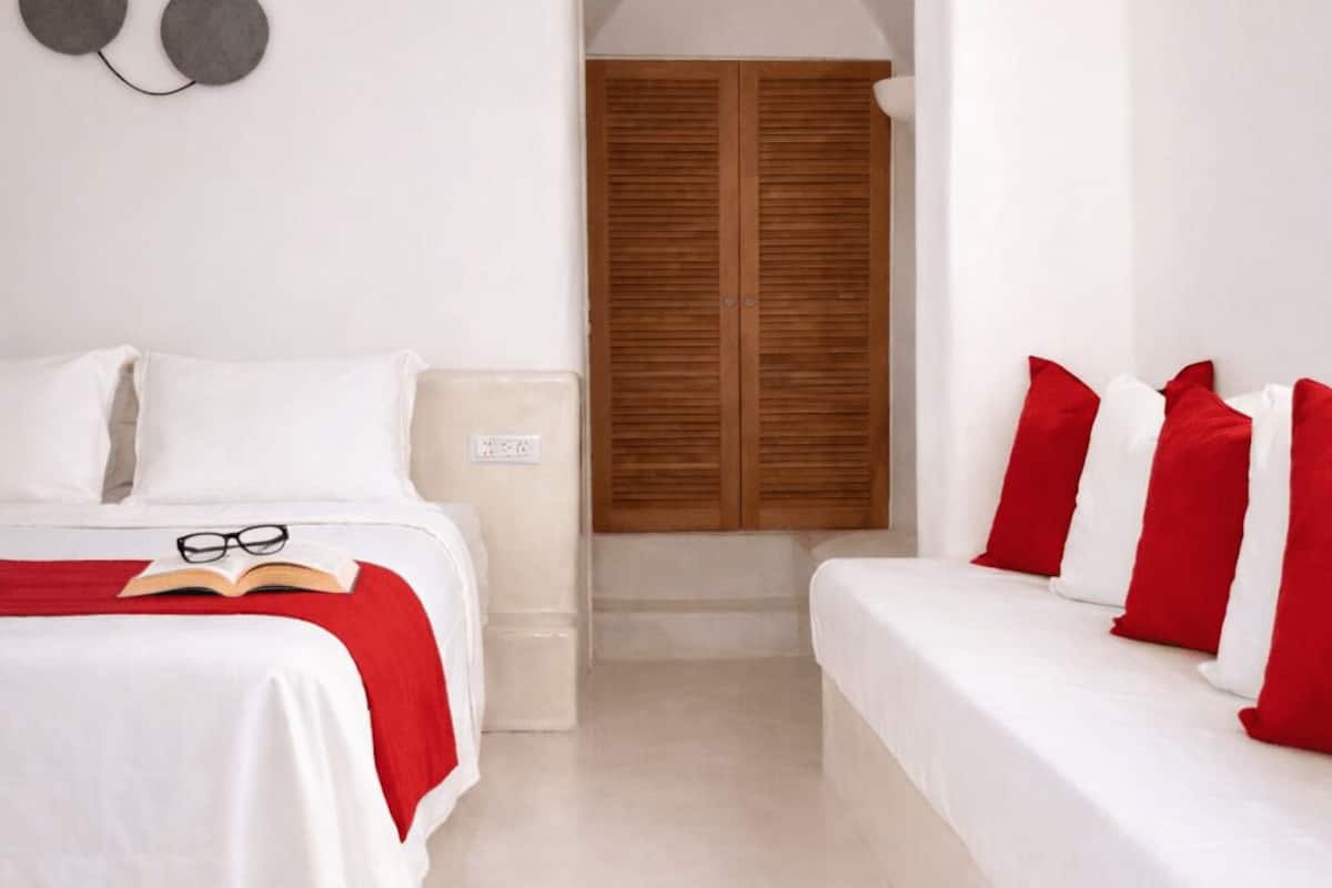 A bedroom with a bed, a bedside table, a wardrobe, and a red and white pillow arrangement.