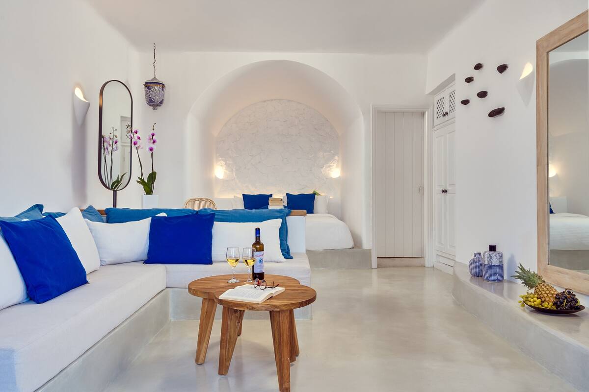 A modern, minimalist living room with a white sofa, blue cushions, a wooden coffee table with a book and wine glasses, and a decorative mirror.
