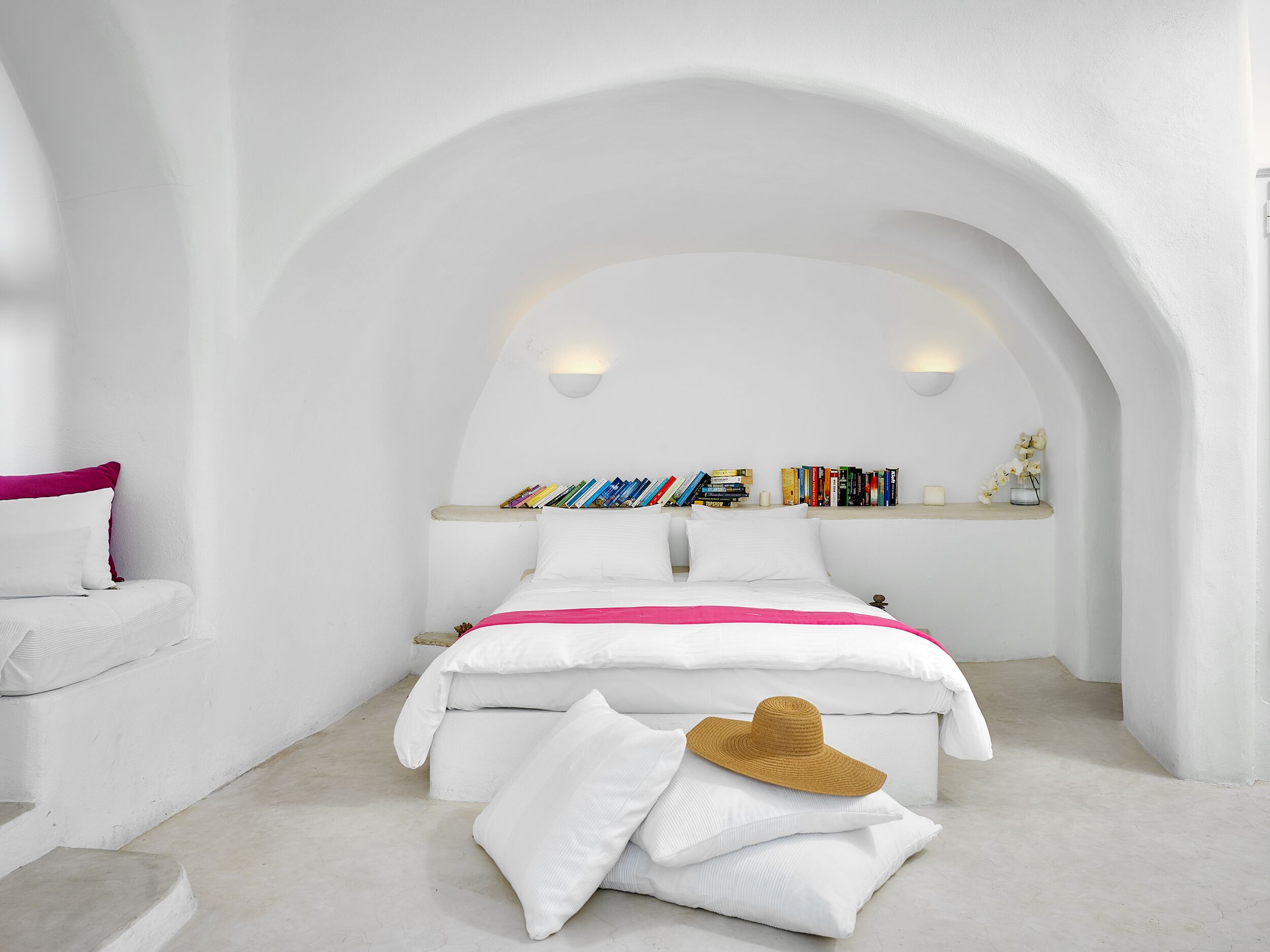 Een slaapkamer in witte tinten met een bed, kussens, een hoed en een boekenplank.