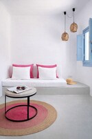 Una habitación minimalista con un sofá blanco, dos almohadas rosas, una mesa redonda con una revista y gafas de sol, una alfombra tejida y una puerta azul.