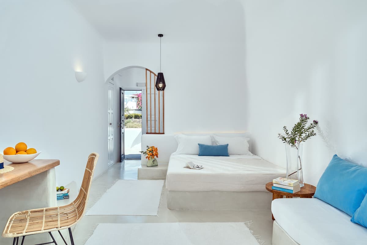 A modern, minimalist bedroom with a white bed, a wooden side table, a wicker chair, and a view of the outdoors.