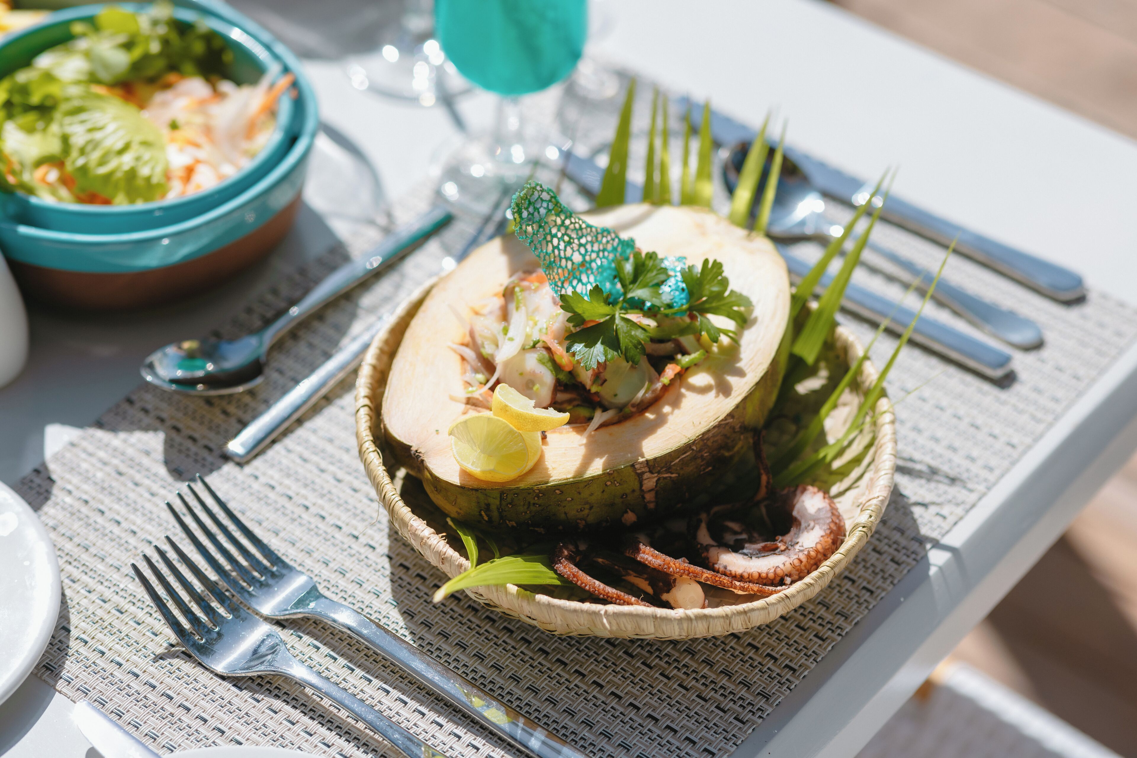 Un plato de mariscos sobre una estera tejida, acompañado de un tenedor y una cuchara, con un vaso de agua y un tazón de ensalada al fondo.