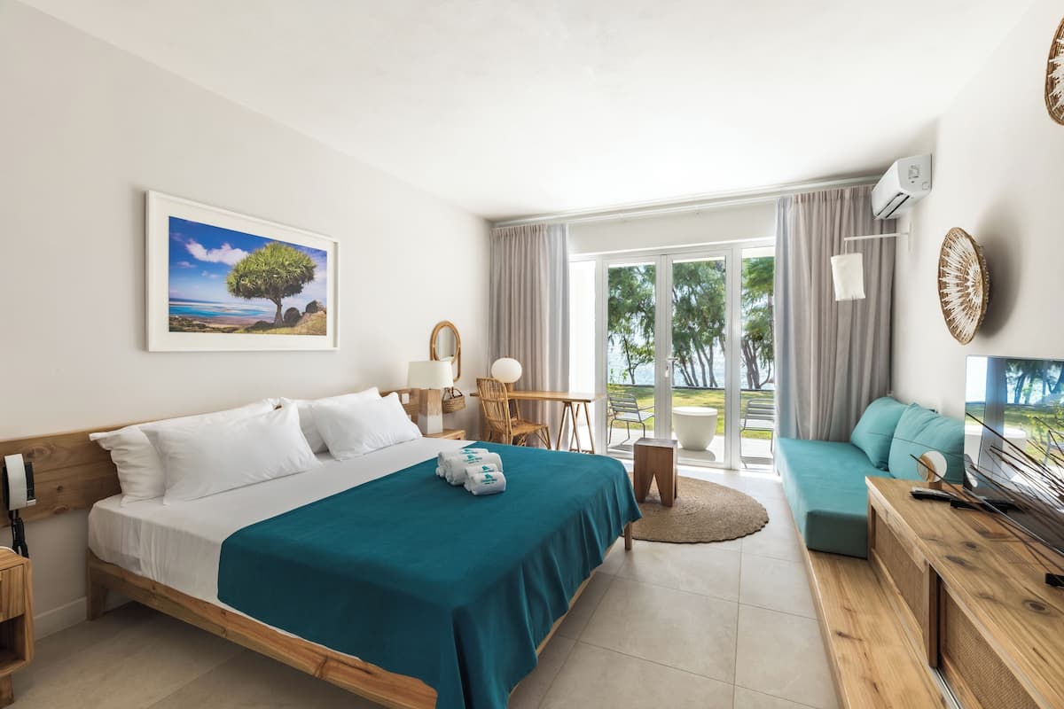 A hotel room with a bed, a sofa, a TV, a desk, and a balcony with a view of trees.
