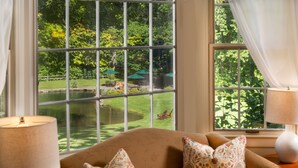 A room with a large window overlooking a pond and greenery, a sofa with patterned cushions, and two lamps on side tables. - Grafton Inn (Grafton)