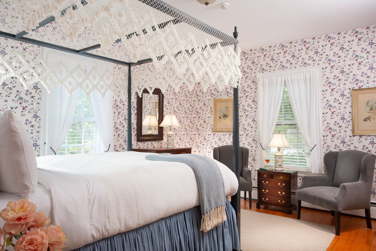 Ein Bett mit Baldachin, Blumenwallpaper, ein Schminktisch mit Spiegel, ein Sessel und ein Fenster mit Vorhängen.