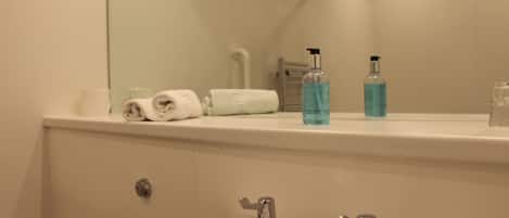 Un baño con lavamanos, dos grifos, un espejo y una encimera con toallas y artículos de aseo.