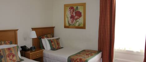 Habitación de hotel con dos camas, una mesita de noche con lámpara, un cuadro en la pared y una ventana con cortinas.