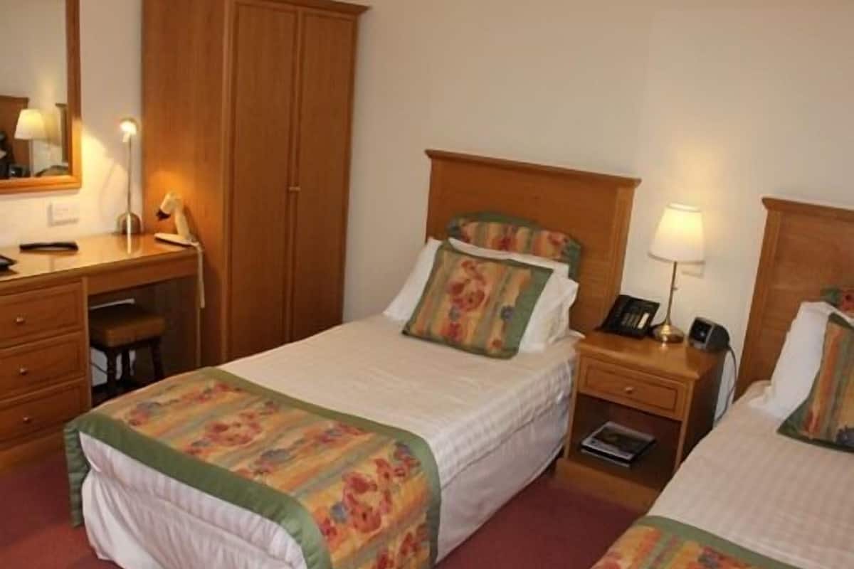 Quarto de hotel com duas camas de solteiro, guarda-roupa de madeira, escrivaninha com cadeira, mesa de cabeceira com abajur e telefone.