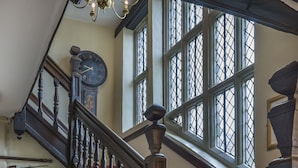 Un magnifique escalier avec une rampe en bois, une grande horloge murale et une fenêtre ornée d’un croisillon en forme de losange.