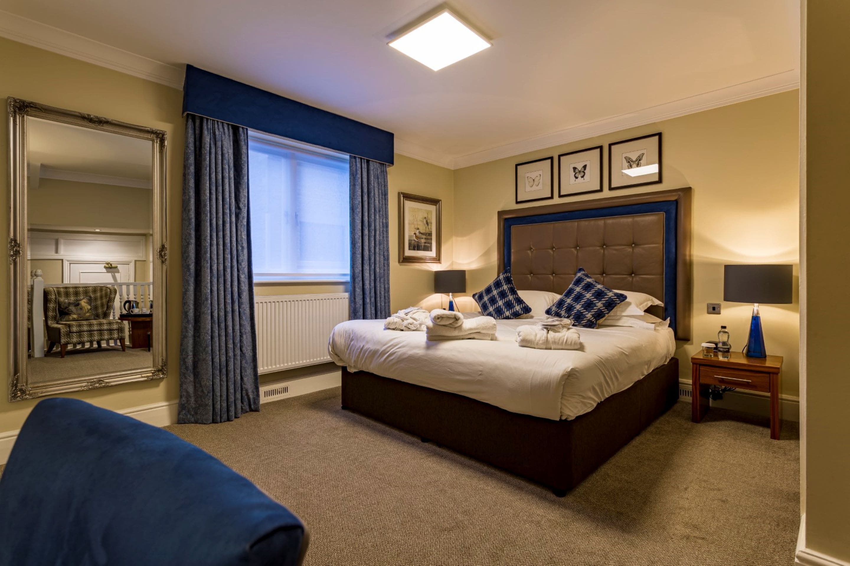 Habitación de hotel bien iluminada con una cama grande, un espejo, una silla y una ventana con cortinas.