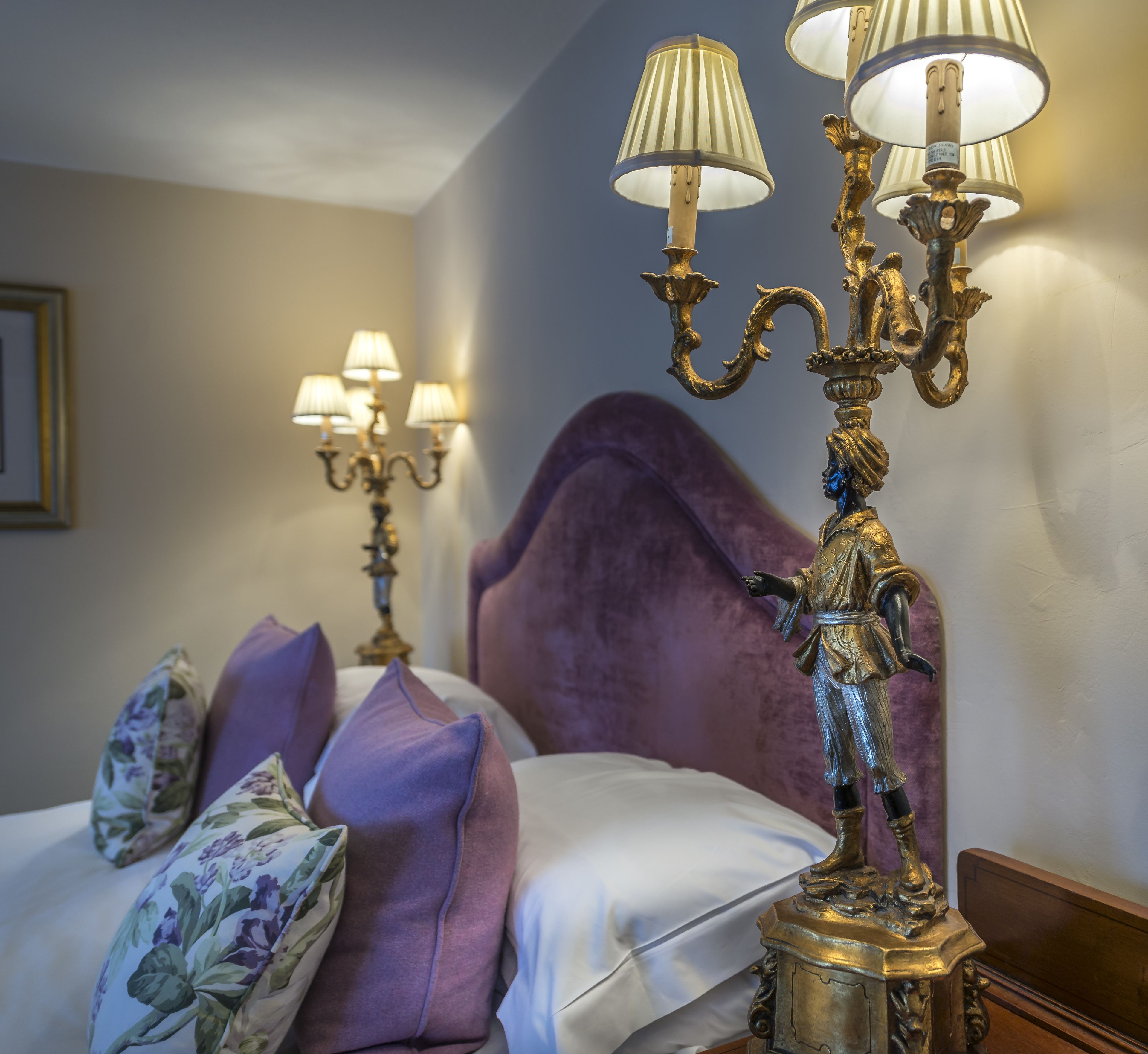 A bed with a purple headboard, a decorative candelabra with multiple lamps, and floral-patterned pillows.