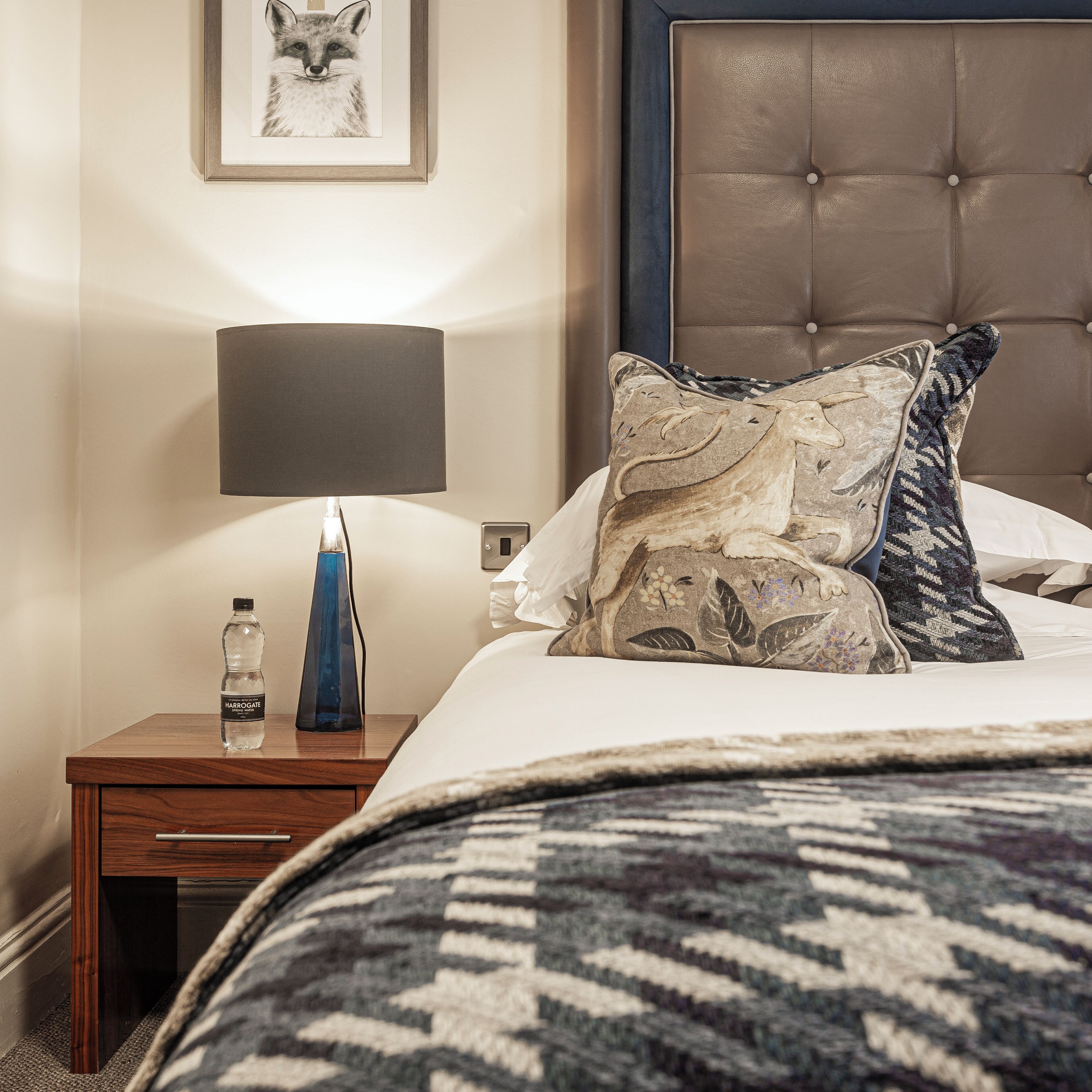 A neatly made bed with a patterned blanket, a bedside lamp, a water bottle, and a framed picture on the nightstand.