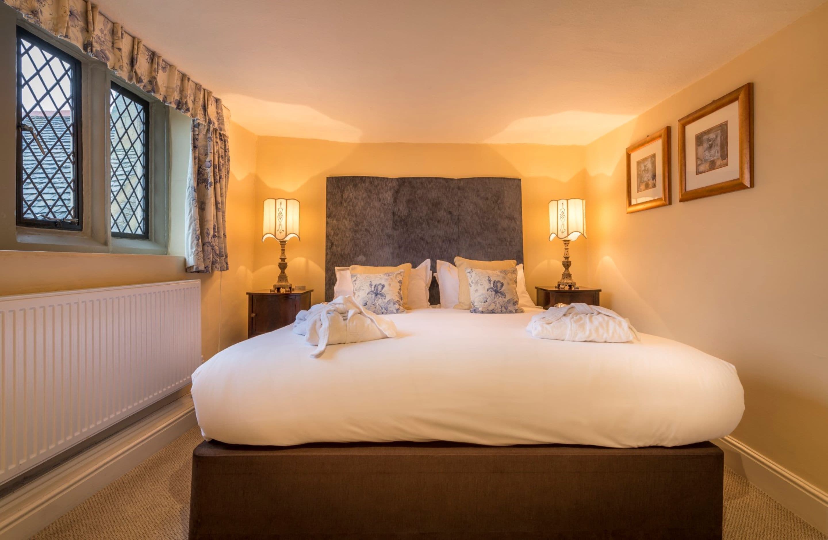Una cama grande con ropa de cama blanca y almohadas estampadas, dos lámparas de noche y un ventanal con cortinas.
