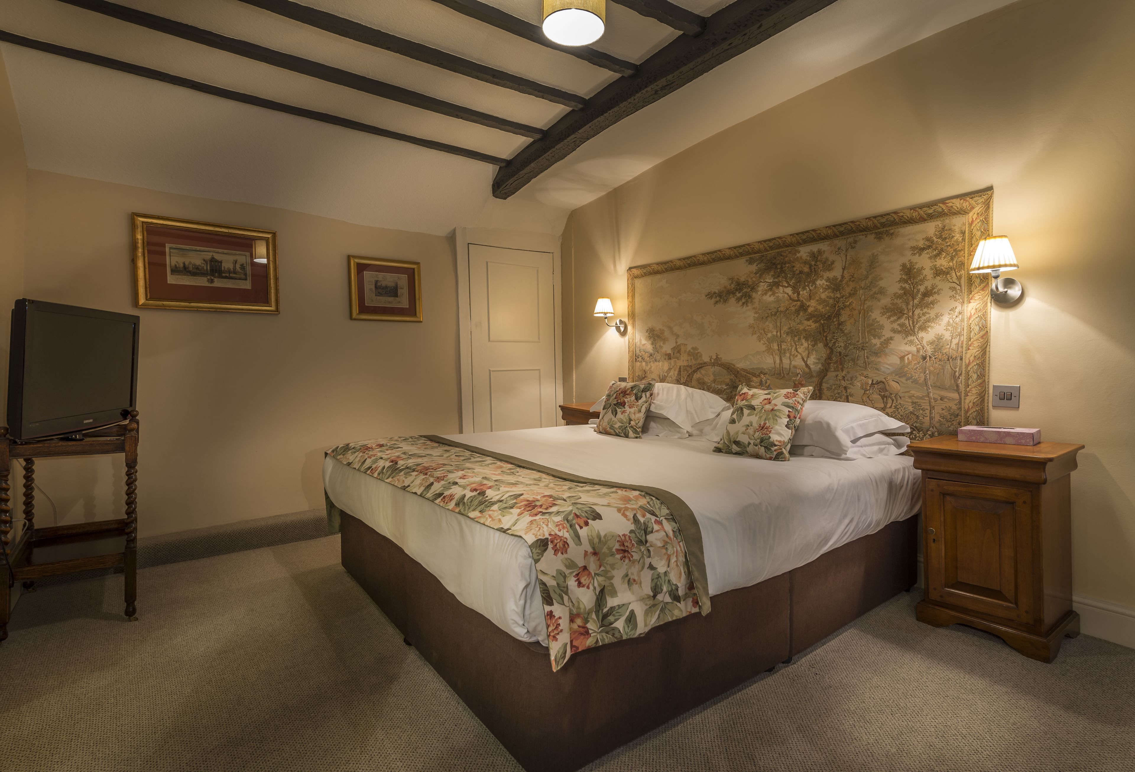 A bedroom with a large bed, wooden headboard, two wall-mounted lamps, a TV on a stand, and framed pictures on the wall.