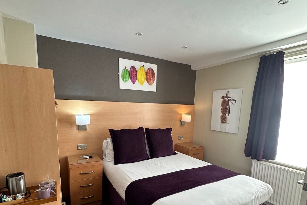 Quarto de hotel com cama, mesas de cabeceira, cômoda, radiador e obras de arte nas paredes.