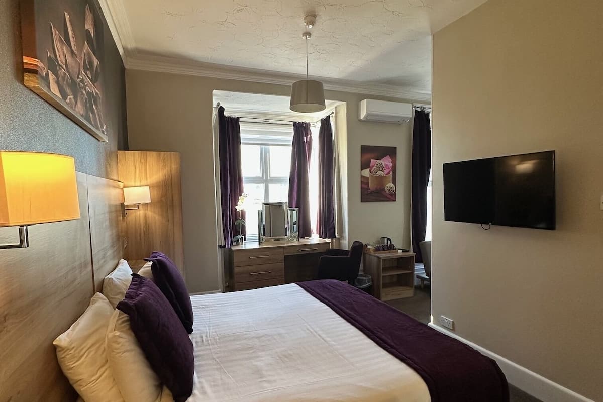 Quarto de hotel com cama, escrivaninha, cadeira, televisão e janela com cortinas.